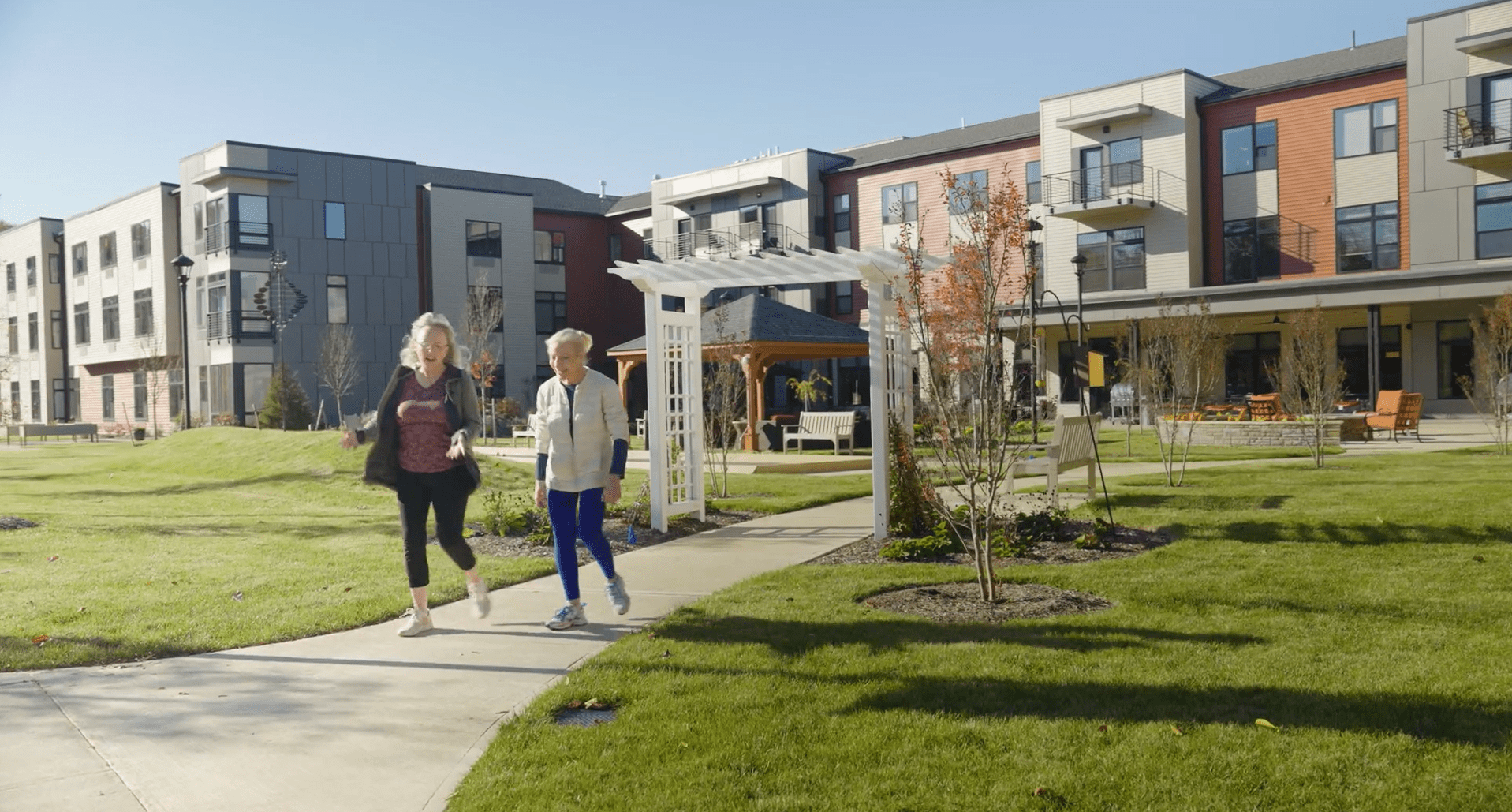 Two seniors walking on a path in the vibrant, green, inviting courtyard with Brightview Wayne behind them. There are small trees and a pergola behind the seniors with a clear blue sky.